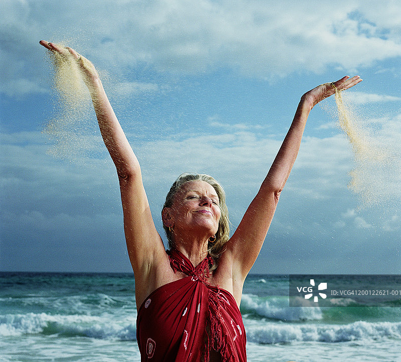 海边的中年妇女，闭着眼睛，举起双臂，沙子从手中落下图片素材