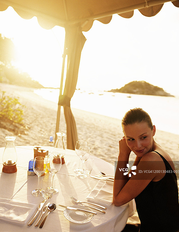 穿黑色连衣裙的女人坐在海滩的餐桌旁，日落时分图片素材