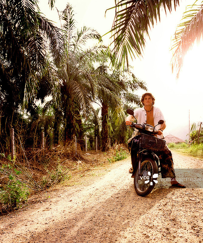 男人坐在棕榈树环绕的土路上的摩托车上图片素材