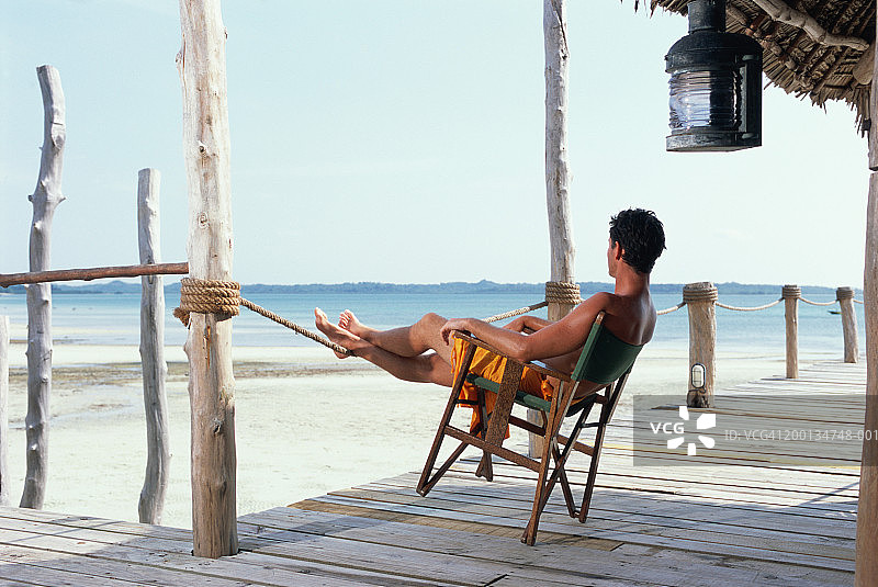 男子向后靠在海滨阳台的椅子上，后视图图片素材
