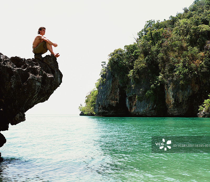 年轻男人坐在岩石上，俯瞰大海，侧视图图片素材
