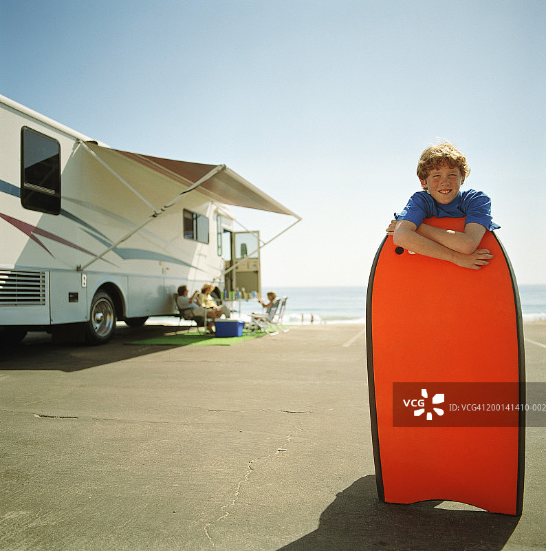 拿着冲浪板的男孩（9-11岁），背景是家庭房车露营图片素材