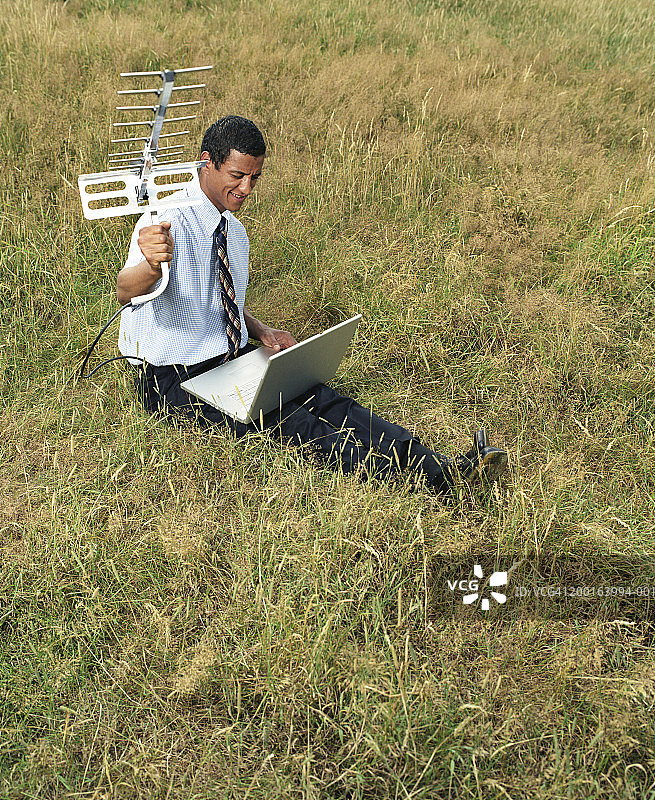 男人坐在田野里，使用笔记本电脑并拿着天线图片素材
