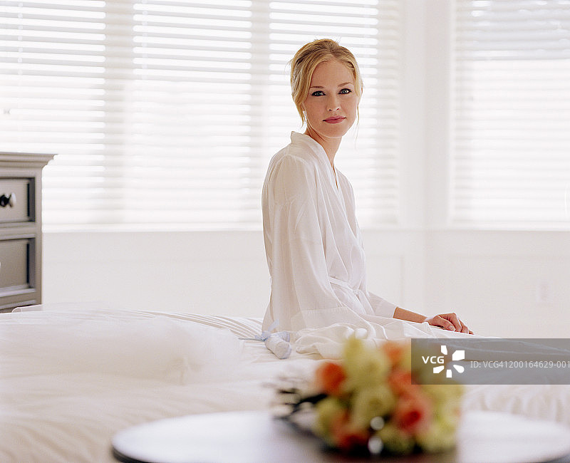 年轻新娘坐在婚纱和面纱旁边的床上，肖像照图片素材