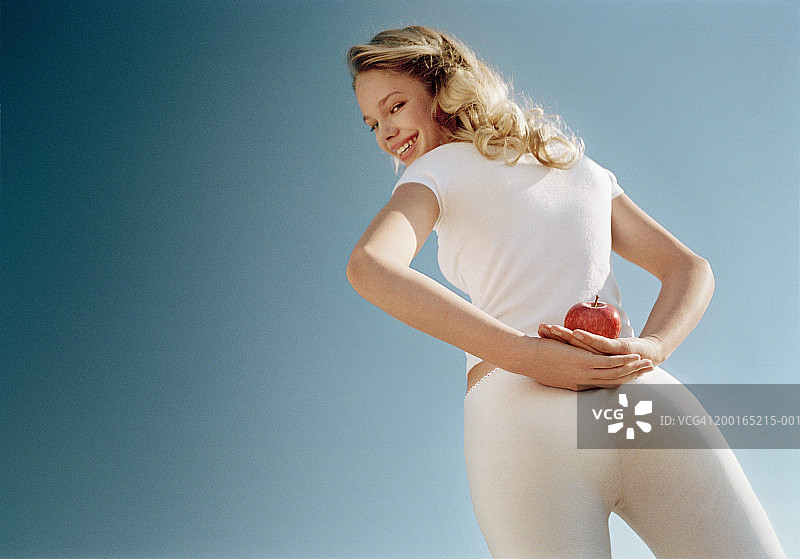 背后藏着苹果，面带微笑的年轻女子肖像，低角度拍摄图片素材