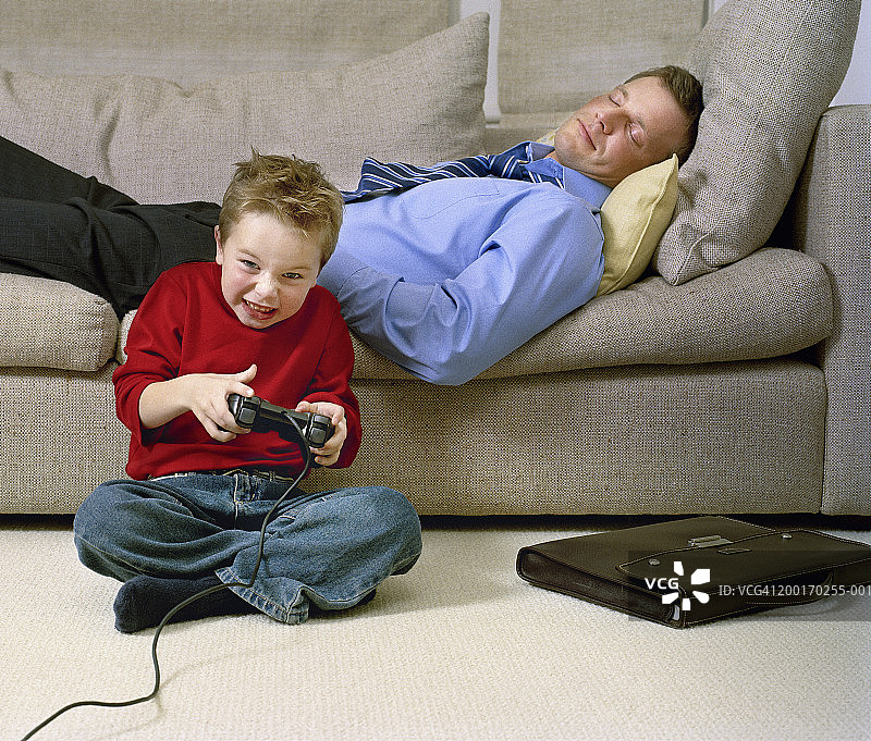 男孩（4-6岁）在玩游戏机，父亲在沙发上睡着了图片素材