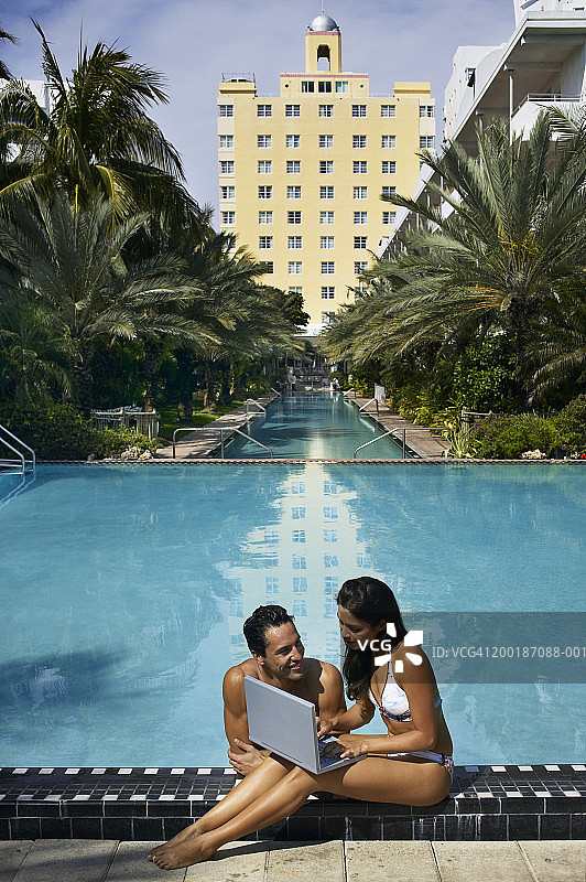 情侣在泳池边使用笔记本电脑，男人对着女人微笑图片素材