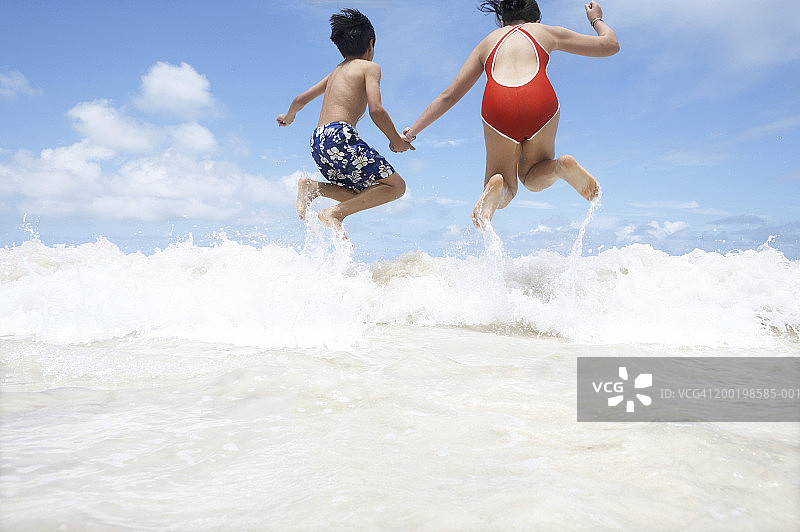 兄妹（9-11岁）手牵手在海浪中跳跃，后视图图片素材
