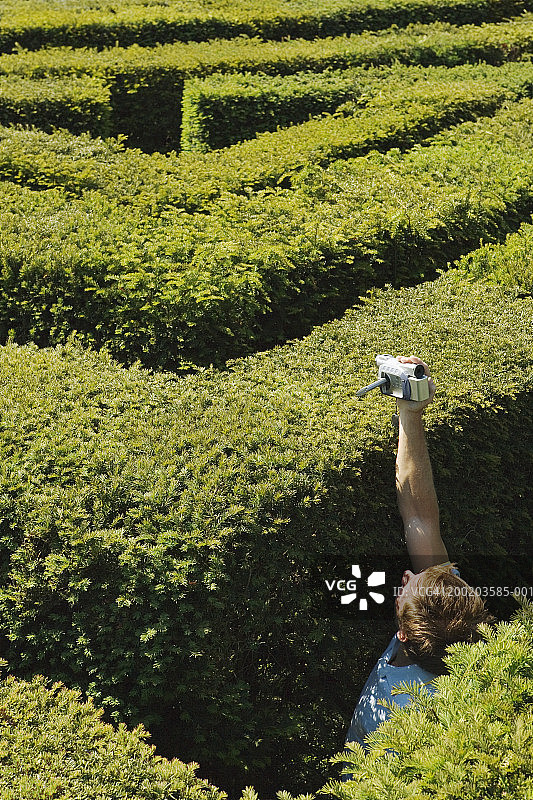 在迷宫中，手持摄像机越过树篱的年轻男人图片素材