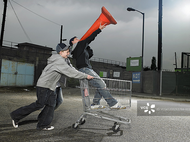 （13-15岁）青少年男孩们推着拿着交通锥的朋友坐在手推车里图片素材