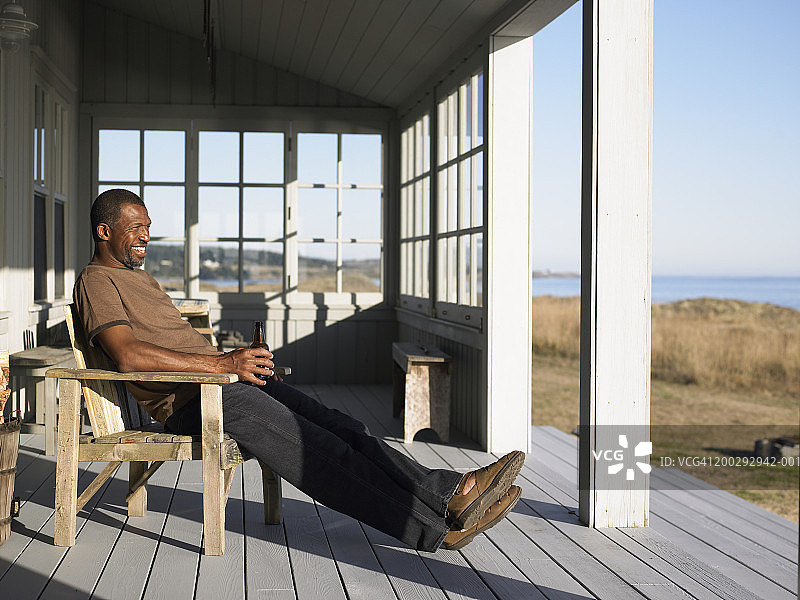 中年男人坐在门廊的扶手椅上喝啤酒，侧视图图片素材