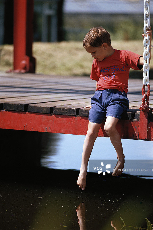 坐在码头上，把脚浸在水里，向下看的4-5岁男孩图片素材