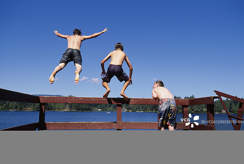 三个男孩在码头上，一个看着朋友们从栏杆跳入湖中（后视图）图片素材