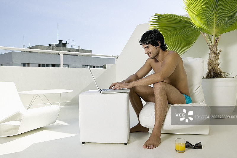 年轻男人穿着泳裤在屋顶使用笔记本电脑，侧面视角图片素材