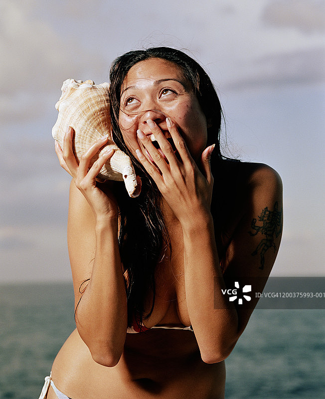 年轻女子身穿比基尼，拿着海螺壳贴在耳朵上，笑着图片素材