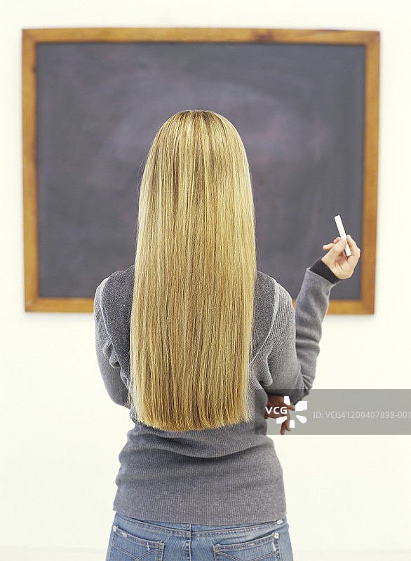 年轻女子看着黑板，手拿粉笔，后视图图片素材
