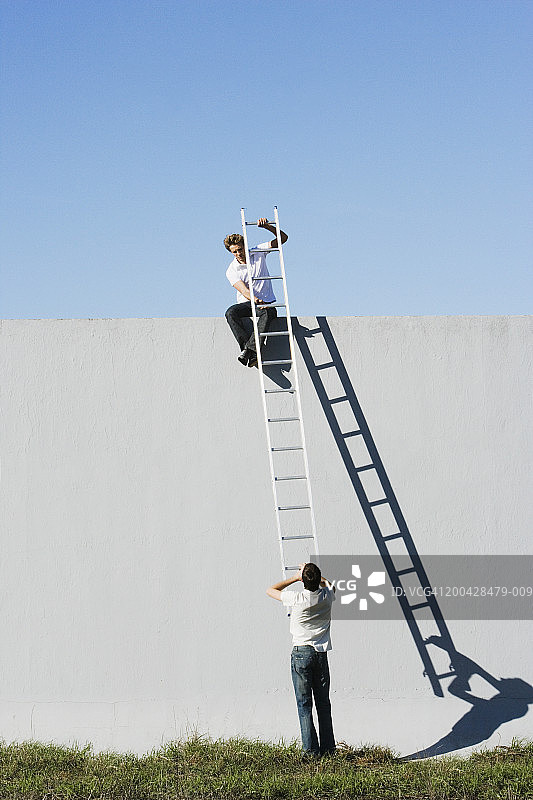 站在墙顶拿着梯子的男人，后视图，地面上有另一个人图片素材