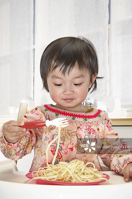 坐在高脚椅上用叉子吃意大利面的女婴（18-24个月）图片素材