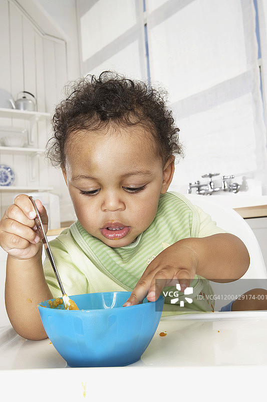 坐在高脚椅上手拿勺子看着碗里的男婴（18-24个月）图片素材