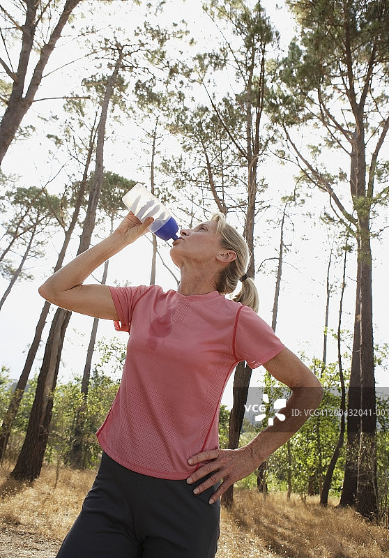 中年妇女在树林里喝运动水，手扶着臀部图片素材