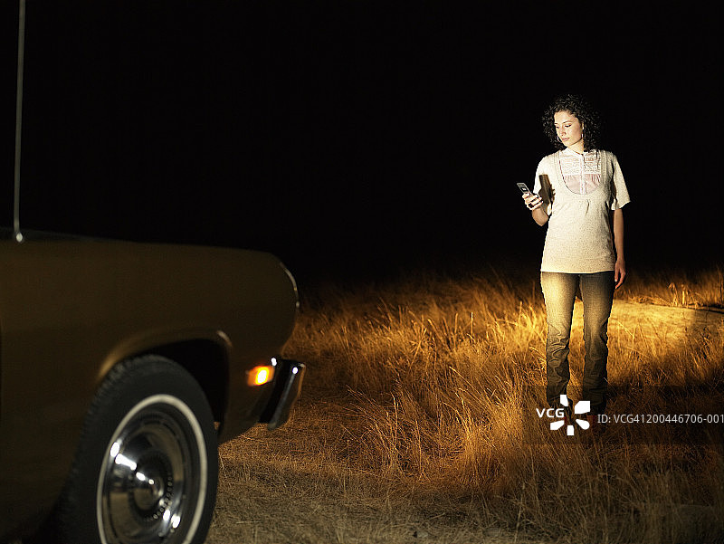 年轻女人在停放的汽车前使用手机，夜晚图片素材