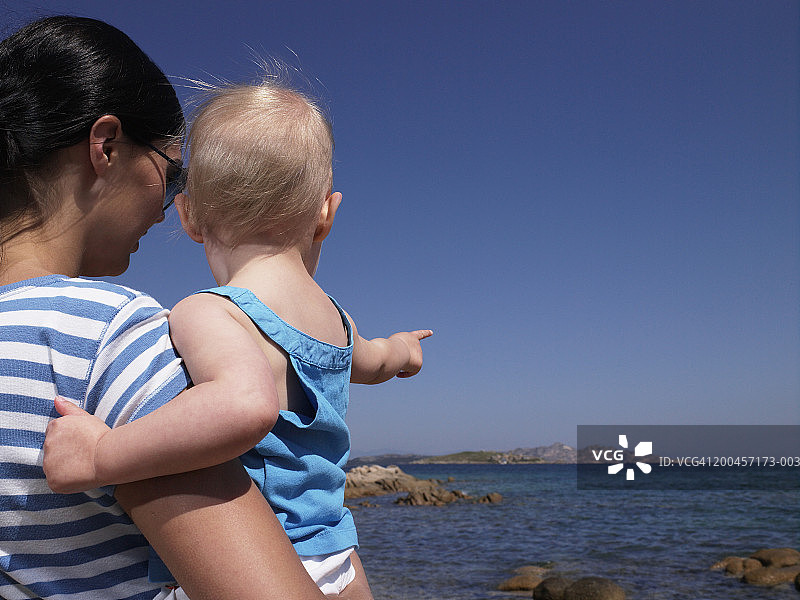 母亲抱着面向大海的女婴（15-18个月），后视图图片素材