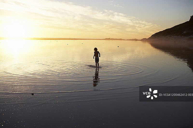 日落时分，4-6岁男孩站在泻湖浅水中（后视图）图片素材