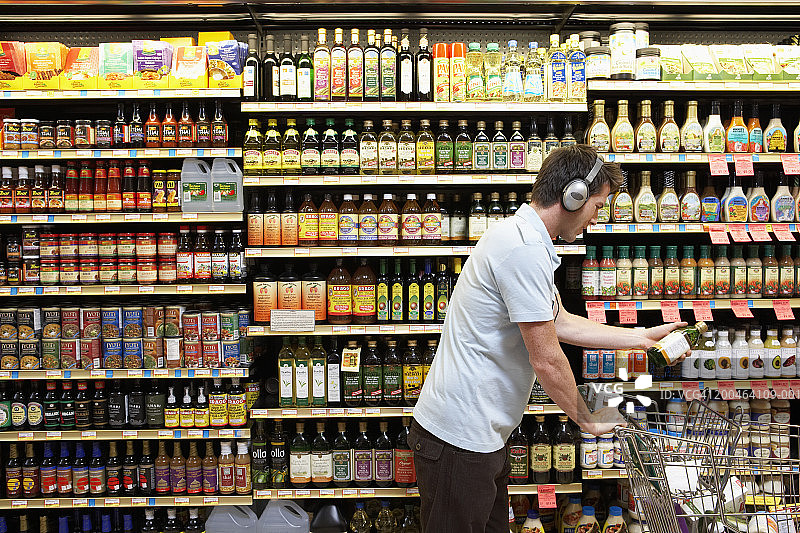戴着耳机的年轻男子将油瓶放入购物车，侧视图图片素材