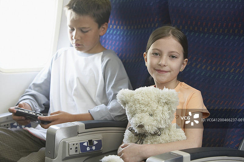 在飞机上抱着泰迪熊的女孩（6-8岁）和玩电子游戏的男孩（9-11岁）图片素材