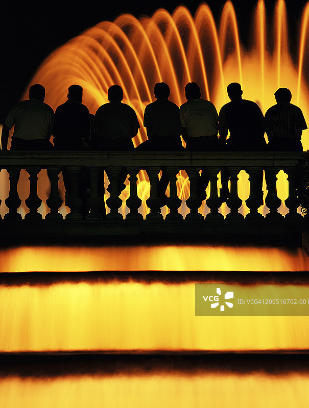 顾客坐在栏杆上，面对着灯光喷泉的剪影图片素材