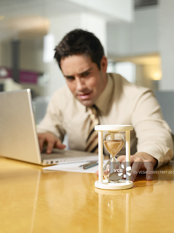 商人坐在笔记本电脑前，看着桌上的沙漏图片素材