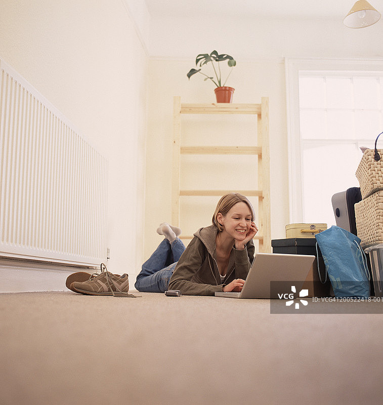 年轻女子躺在地板上，身旁有一堆物品，使用笔记本电脑图片素材