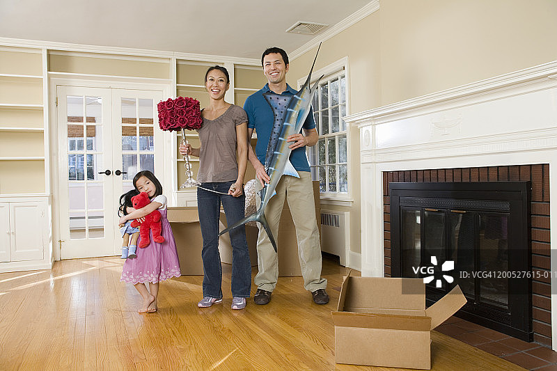 夫妇与年幼的女儿(4-5岁)在客厅里微笑合影，手持个人物品，家庭照图片素材