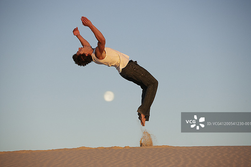 年轻男人在沙漠中表演后空翻，侧视图，黄昏图片素材