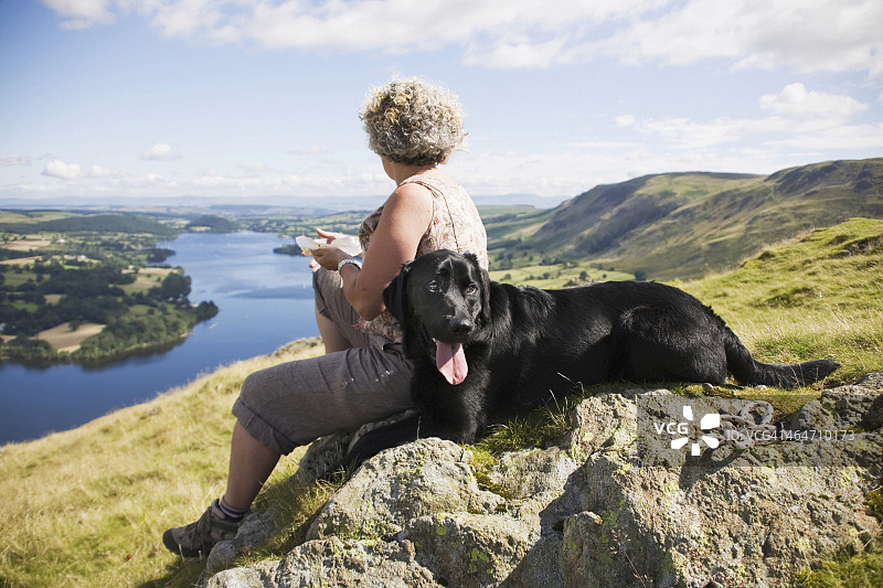 一位女士和她的狗坐在一块岩石上，俯瞰着一个大湖和风景图片素材