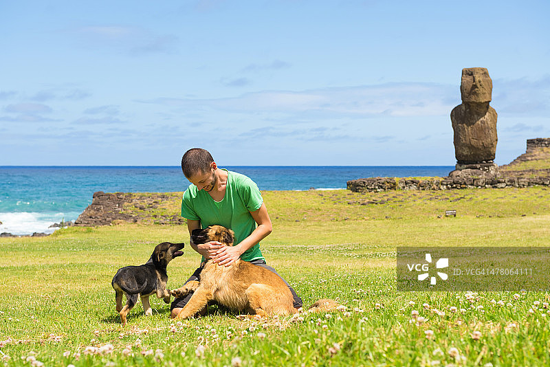 男人在草地上和两只狗玩耍，背景是摩艾石像图片素材