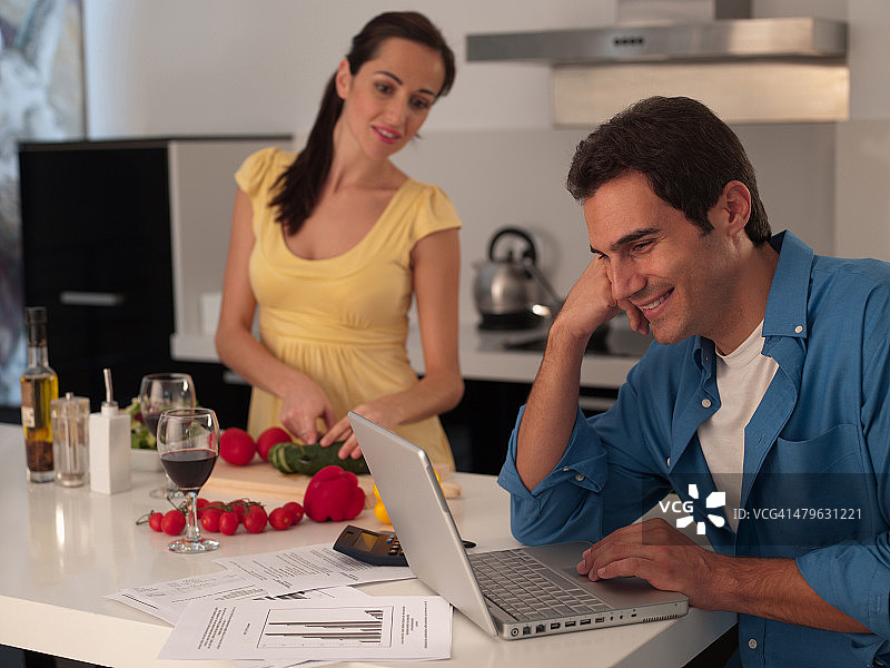 在现代厨房里，男人用笔记本电脑工作，女人准备饭菜图片素材