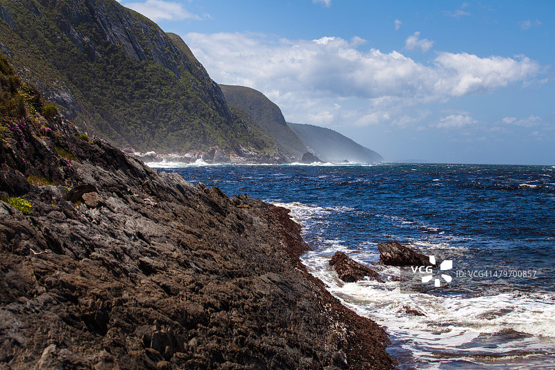 南非西开普省花园大道齐齐卡马国家公园的岩石海岸和山脉被蔚蓝的印度洋冲刷图片素材