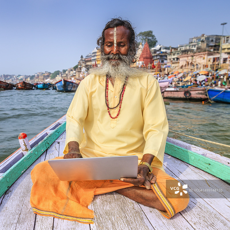 瓦拉纳西，在恒河圣河的船上使用笔记本电脑的印度圣人图片素材