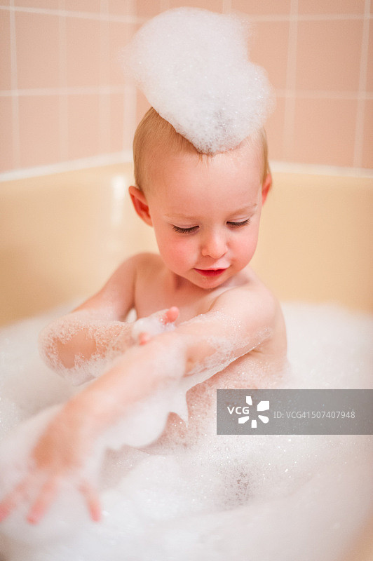 一个快乐的金发幼儿女孩坐在满是泡泡的浴缸里，自己洗澡图片素材