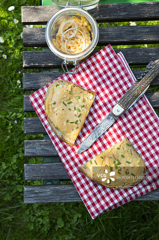 花园桌上放着两片撒有细香葱、布、刀和蜜饯罐的奥巴兹达面包片图片素材