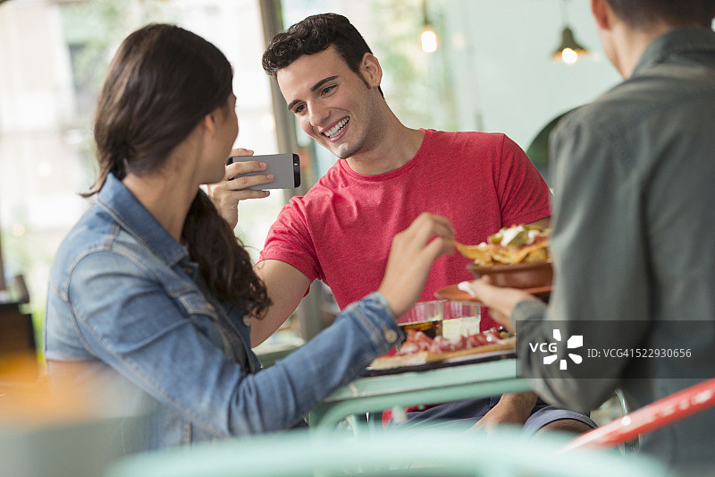 在咖啡馆的桌子旁用智能手机给女人拍照的微笑男人图片素材