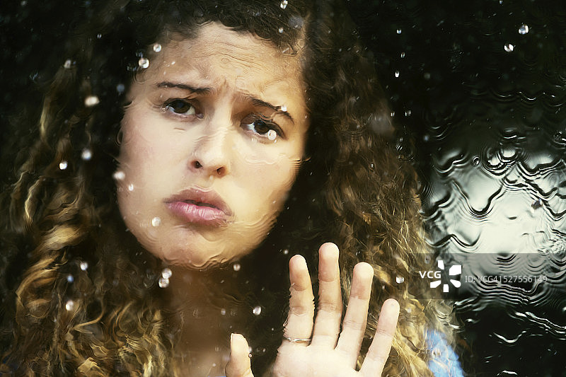 悲伤的女人隔着雨点斑驳的窗户向外看。压抑的天气，沮丧的女孩图片素材