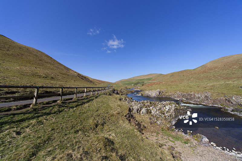 沿着道路的木栅栏，小溪穿过山丘；英国诺森伯兰郡切维厄特图片素材