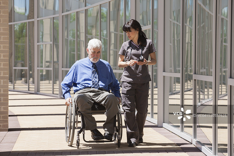 坐在轮椅上使用智能手机，由护士陪同的患有肌营养不良症和糖尿病的男人图片素材