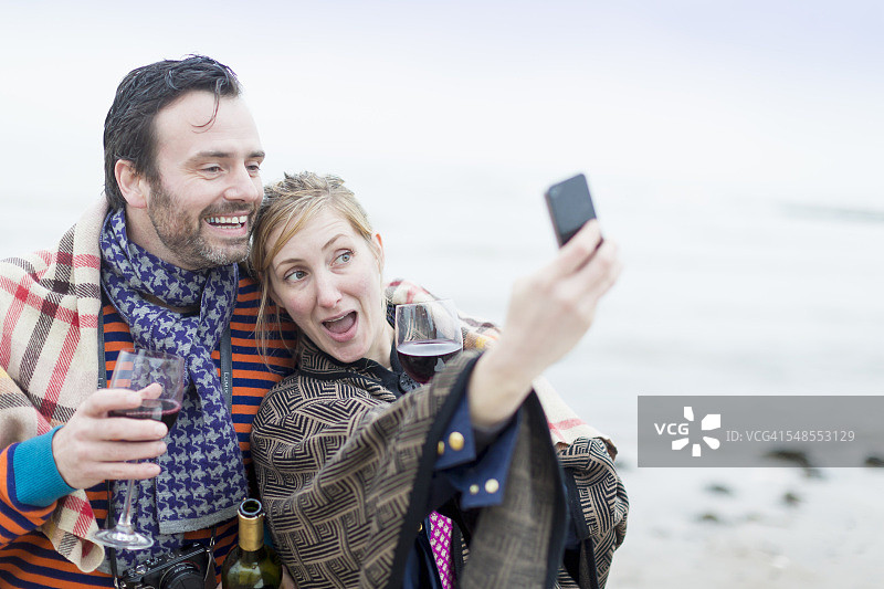 情侣在海滩上喝着葡萄酒，用智能手机自拍图片素材