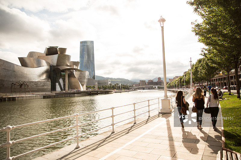 在西班牙毕尔巴鄂古根海姆博物馆附近海滨漫步的三名女性游客图片素材