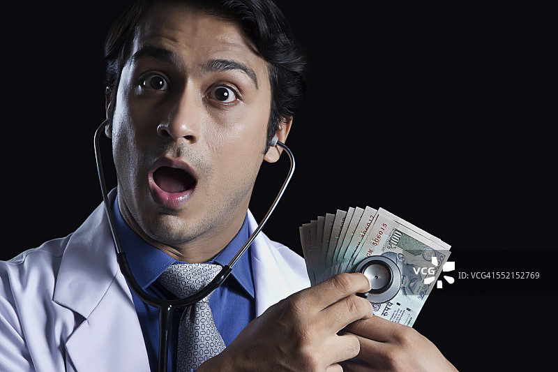 震惊的男医生用听诊器检查印度货币，黑色背景图片素材