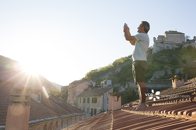 男人在瓷砖屋顶上用智能手机拍照图片素材
