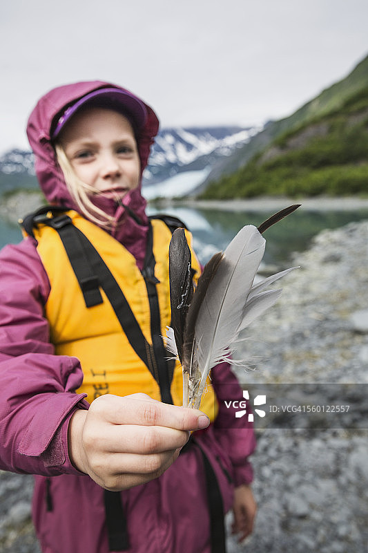 穿着黄色救生衣,展示一些鸟类羽毛,站在舒普冰川和楚加奇山脉前的年轻女孩,舒普湾州立海洋公园,威廉王子湾图片素材